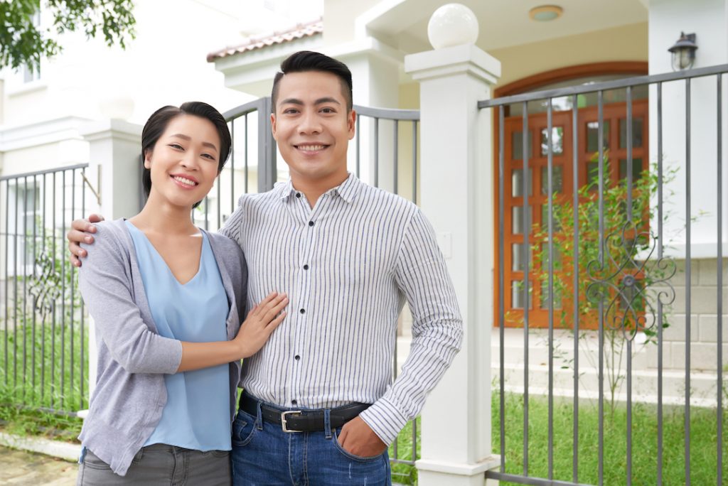 Couple posing for photo infront of house | Featured image for Love and Mortgages: Buying a Home with a Partner blog